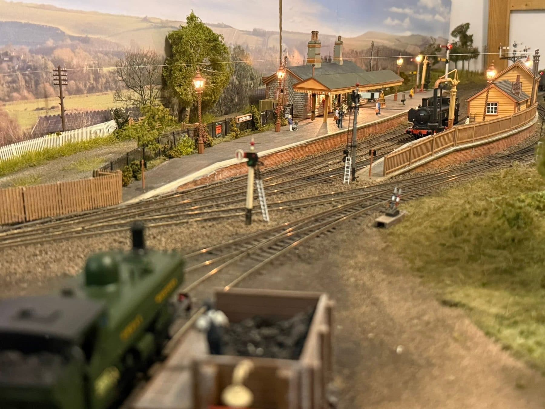 Buckleigh station from the end of the loop: the stone station building and lit platform lamps, a pannier tank at the platform and semaphore signals in the foreground