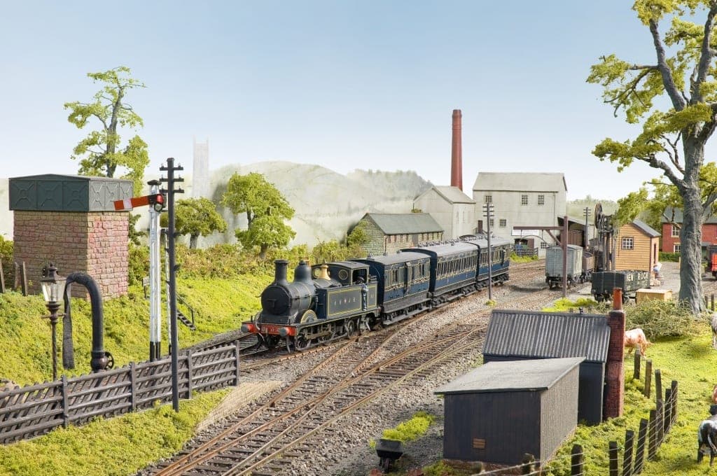 An S&DJR tank engine brings blue coaches into Cheddar past a brick water tower and signal, with the mill chimney and the hills behind