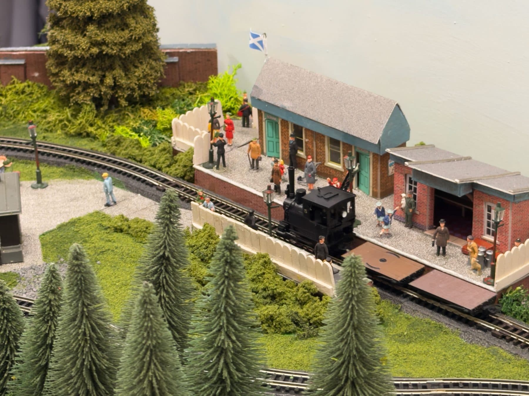 A small black tank engine at the platform of Glen McHoo Halt, with passengers waiting and a Saltire flying above the station