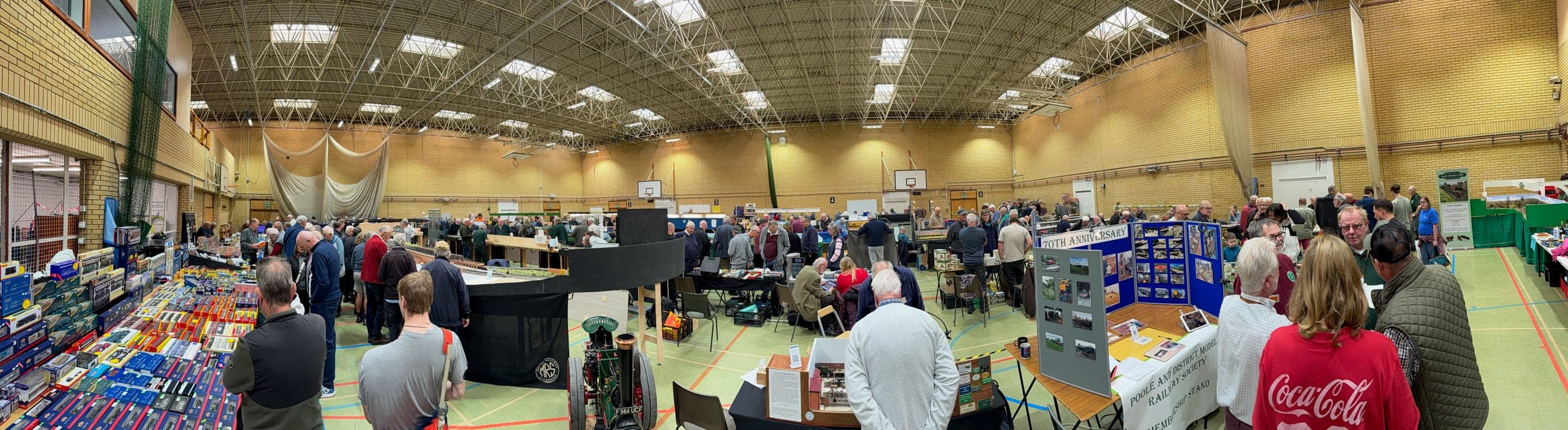 A panorama of the busy hall at the 2026 show: layouts, trade stands and visitors under a roof of skylights, with the society's 70th anniversary display boards on the right