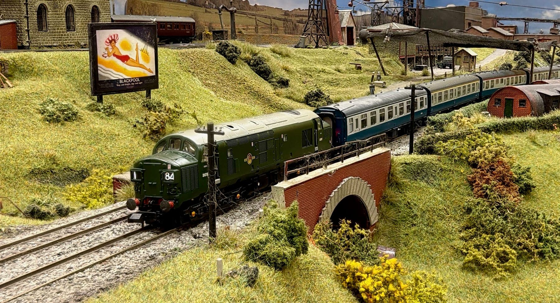 A green diesel leads a train of blue and grey coaches over the brick underbridge, with the colliery headgear and ropeway behind