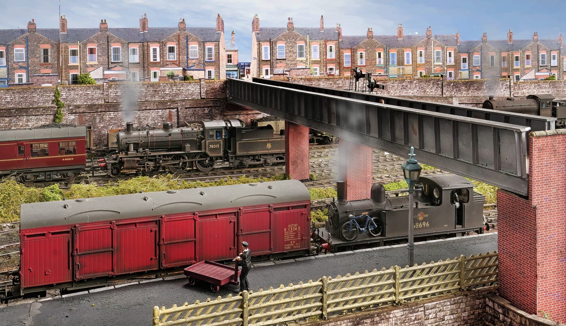 BR tank engines pass beneath a plate girder bridge on brick piers, with a red parcels van in front and terraced houses above
