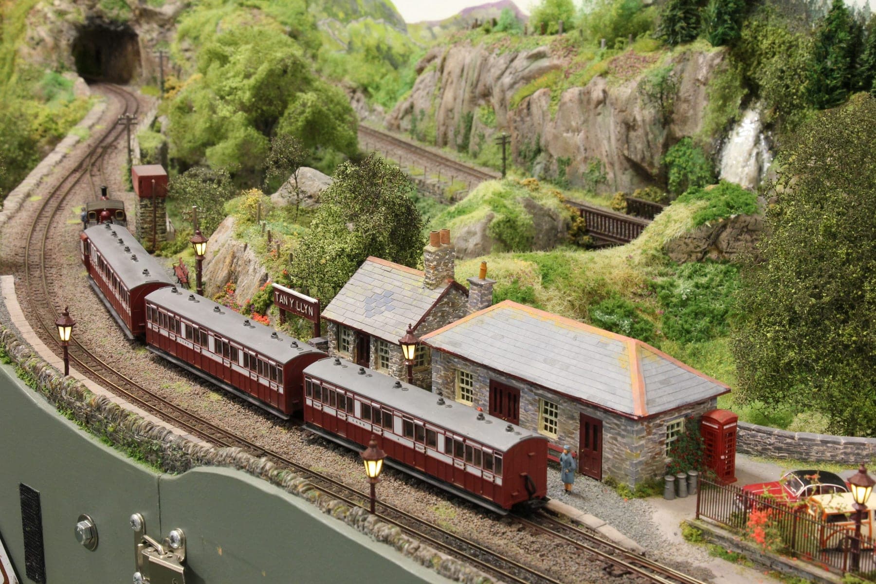 A train of maroon narrow gauge coaches at Tan-y-Llyn's stone station, below wooded crags, a waterfall and a tunnel mouth